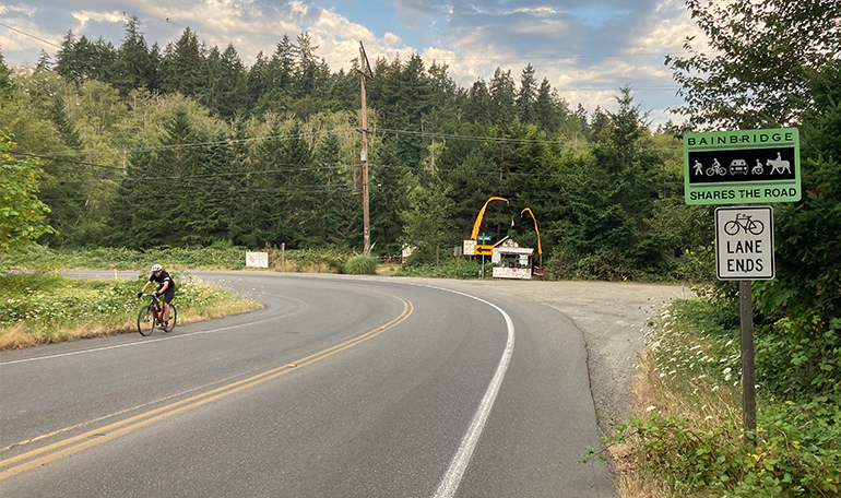Two lane road in rural setting, with bicyclist traveling one direction and Share The Road sign on post visible on other side